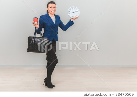 Young teacher in formal suit with bag, holding a white clock, and cup of coffee, setting her teeth Young teacher in formal suit with bag, holding a white clock, and cup of coffee, setting her teeth 58470509