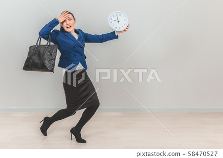 Young teacher in formal suit with bag, holding a white clock, running because she is late. 58470527