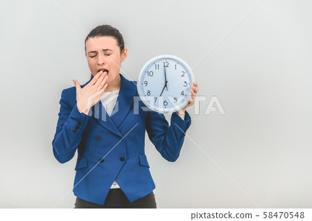 Sleepy and tired young teacher in formal suit is yawning, covering mouth by hand, holding a clock. 58470548