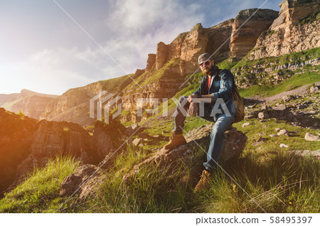 Close-up of a bearded man in jeans clothes in sunglasses and a cap with a backpack sitting at the 58495397