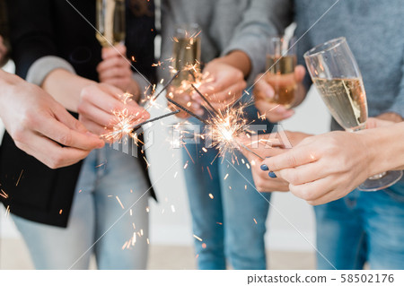 Hands of young friends holding burning bengal lights and flutes of champagne Hands of young friends holding burning bengal lights and flutes of champagne 58502176