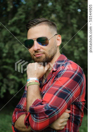 Outdoors portrait of young bearded man with sunglasses looking at the camera in the park on trees Outdoors portrait of young bearded man with sunglasses looking at the camera in the park on trees 58544905