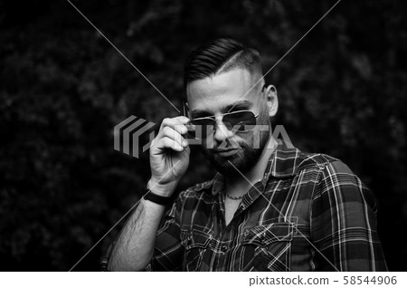 Black and white portrait of young bearded man with sunglasses looking at the camera outdoors in the Black and white portrait of young bearded man with sunglasses looking at the camera outdoors in the 58544906