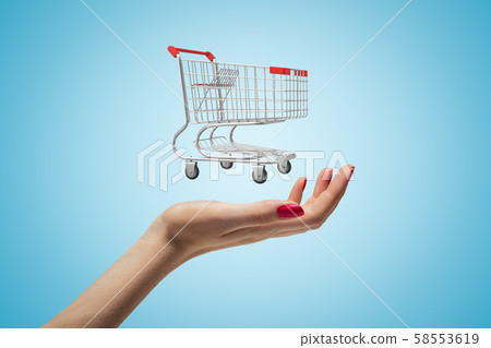 Side closeup of woman's hand facing up and levitating small supermarket trolley on light blue 58553619