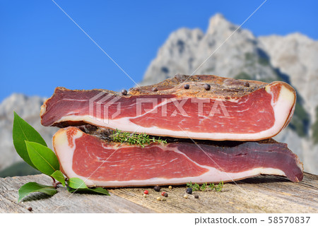 Typical South Tyrolean bacon divided into two halves lying on a rustic table in front of the alps Typical South Tyrolean bacon divided into two halves lying on a rustic table in front of the alps 58570837