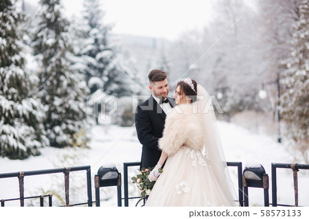 Elegant bride in white fur coat stand with handsome bearded groom in front of snow covered pine 58573133