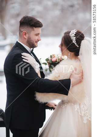 Elegant bride in white fur coat stand with handsome bearded groom in front of snow covered trees 58573156