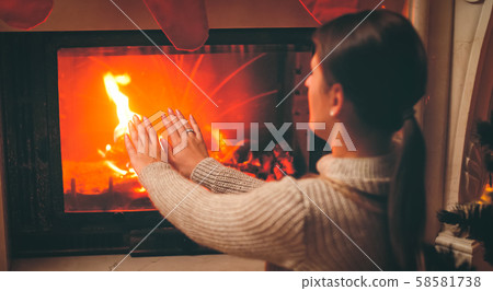 Toned image of beautiful woman feeling cold sitting by the burning fireplace at living room 58581738