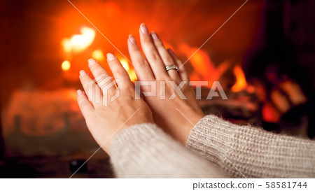 Closeup image of young woman in sweater warming her hands at burning fireplace at house 58581744