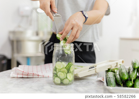 Woman hands with knife putting cucumber into glass jar in kitchen table. Woman hands with knife putting cucumber into glass jar in kitchen table. 58582266