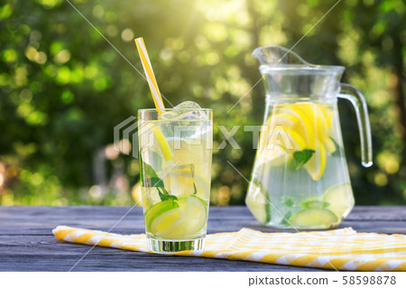 Lemonade with ice in glass with straw and jug on wooden table Lemonade with ice in glass with straw and jug on wooden table 58598878