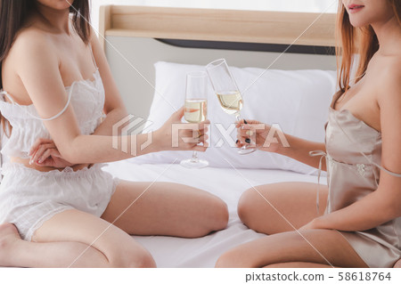 Women in pajamas sit on white bed clink champagne 58618764