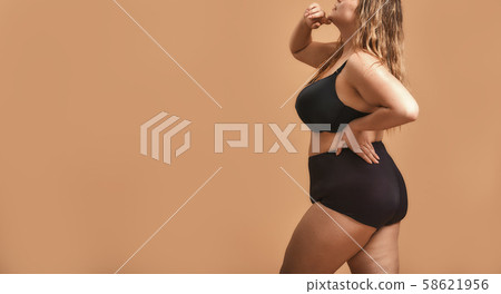 Side view of plus size sexy model in black lingerie standing against brown wall in studio 58621956