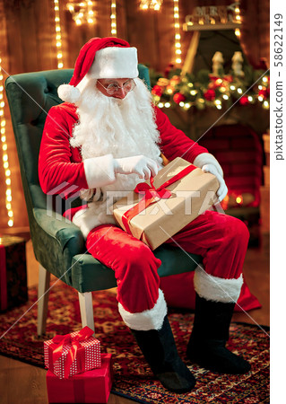 Photo of happy Santa Claus in eyeglasses sitting on chair, hold gift box Photo of happy Santa Claus in eyeglasses sitting on chair, hold gift box 58622149