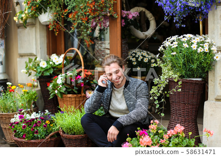 Young caucasian boy talking by smartphone and sitting at flower shop near bouquets in baskets. Young caucasian boy talking by smartphone and sitting at flower shop near bouquets in baskets. 58631471