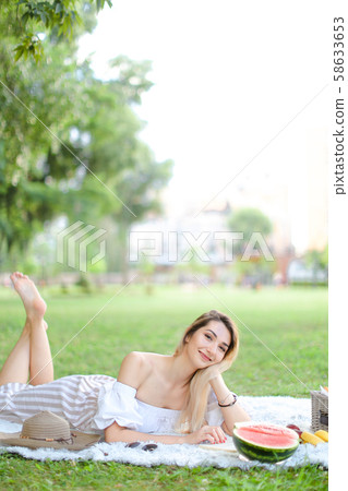 Young girl reading book and lying on plaid near watermelon and hat, grass in background. 58633653