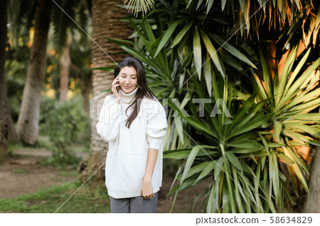 Japanese woman in tropical park talking by smartphone near palm. Japanese woman in tropical park talking by smartphone near palm. 58634829