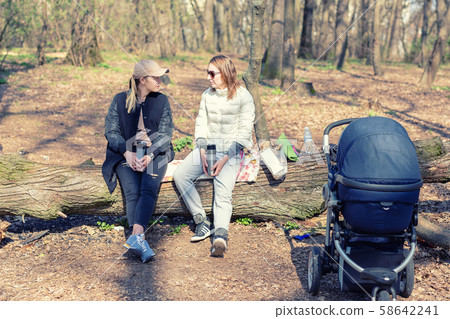 Two women girlfriends sitting on wooden log at forest glade while walking with baby stroller. Young Two women girlfriends sitting on wooden log at forest glade while walking with baby stroller. Young 58642241