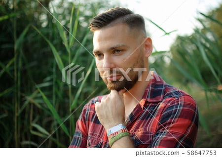 Brutal bearded young man in red shirt is standing outdoors on green stems background. Hipster style Brutal bearded young man in red shirt is standing outdoors on green stems background. Hipster style 58647753
