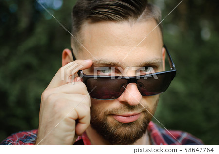 Outdoors portrait of young bearded man with sunglasses looking at the camera in the park on trees Outdoors portrait of young bearded man with sunglasses looking at the camera in the park on trees 58647764