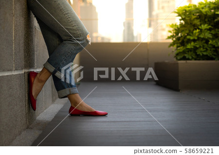 Crop shot of an attractive woman\'s legs in smart casual outfit,\
jeans and ruby red flat shoes, she stand and lean against the wall\
on building rooftop in the evening. Modern city leisure\
lifestyle.