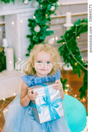 Cute girl with blond hair in a white ball gown by the Christmas tree in holiday dresses with gifts and silver confetti. Christmas mood 58664575