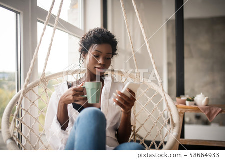 Curly woman sitting and drinking coffee while reading e-mail 58664933