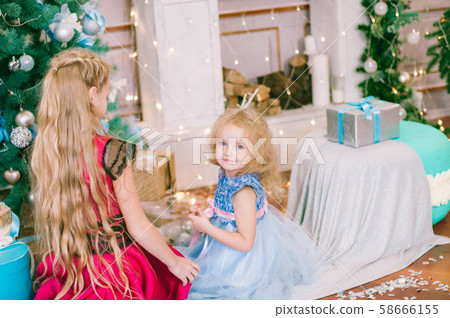 Cute girls  with blond hair in a white ball gown by the Christmas tree in holiday dresses with gifts and silver confetti. Christmas mood 58666155
