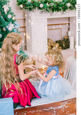 Cute girls with blond hair in a white ball gown by the Christmas tree in holiday dresses with gifts and silver confetti. Christmas mood Cute girls with blond hair in a white ball gown by the Christmas tree in holiday dresses with gifts and silver confetti. Christmas mood 58666157
