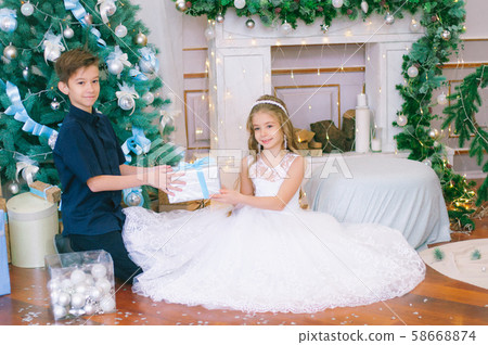 Children, a boy and a girl in a white ball gown near a Christmas tree in holiday dresses with gifts and silver confetti. Christmas mood Children, a boy and a girl in a white ball gown near a Christmas tree in holiday dresses with gifts and silver confetti. Christmas mood 58668874