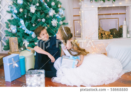 Children, a boy and a girl in a white ball gown near a Christmas tree in holiday dresses with gifts and silver confetti. Christmas mood Children, a boy and a girl in a white ball gown near a Christmas tree in holiday dresses with gifts and silver confetti. Christmas mood 58668880