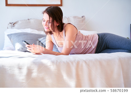 Image of young brunette with phone in her hands lying on bed Image of young brunette with phone in her hands lying on bed 58670199