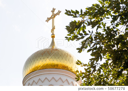 Bottom view of shiny christian church dome with cross on top against leaves and sky 58677278