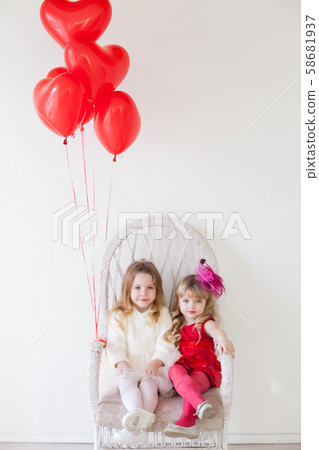 Two girls sit on a chair with red holiday balloons Two girls sit on a chair with red holiday balloons 58681937