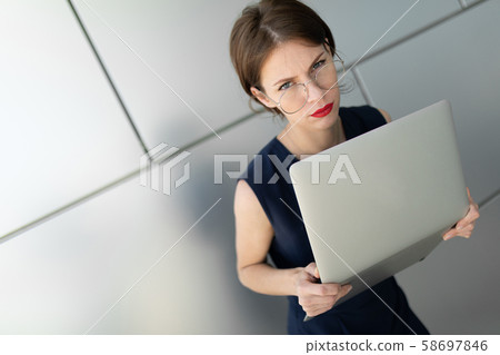 businesswoman holds laptop in her hands, looks in disbelief at the camera, portrait against a gray 58697846
