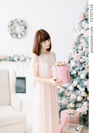 Smiling pretty young woman in light orange pink dress holding christmas present box and standing in Smiling pretty young woman in light orange pink dress holding christmas present box and standing in 58707418