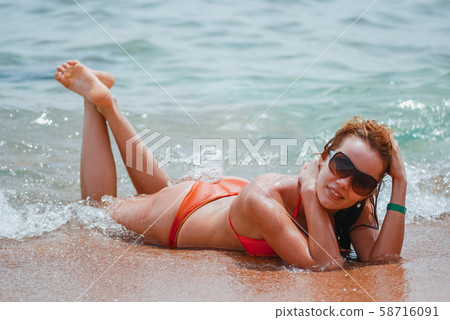 Laughing and happy girl in an orange swimsuit is lying on the wet sea beach and enjoying the waves Laughing and happy girl in an orange swimsuit is lying on the wet sea beach and enjoying the waves 58716091