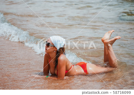 Happy girl in an orange swimsuit is lying on the wet sea beach and enjoying the waves washing the Happy girl in an orange swimsuit is lying on the wet sea beach and enjoying the waves washing the 58716099