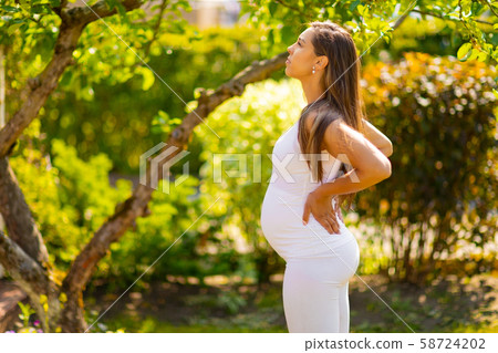 Exhausted pregnant woman standing in garden holding hands on her back Exhausted pregnant woman standing in garden holding hands on her back 58724202