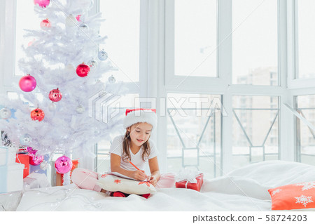 Little girl in Santa hat writes letter to Santa near christmas tree in the cold winter morning. Little girl in Santa hat writes letter to Santa near christmas tree in the cold winter morning. 58724203