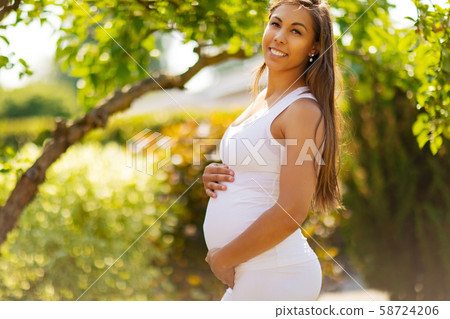 Smiling pregnant woman standing in garden holding hands on belly Smiling pregnant woman standing in garden holding hands on belly 58724206