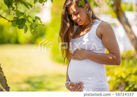 Smiling pregnant woman standing in garden holding hands on belly Smiling pregnant woman standing in garden holding hands on belly 58724337