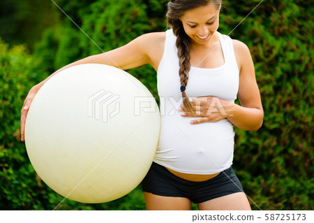 Pregnant Woman Touching Belly While Holding Fitness Ball In Park 58725173