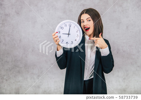 Pretty office female employee standing, pointing her finger at the clock in her hand. 58733739