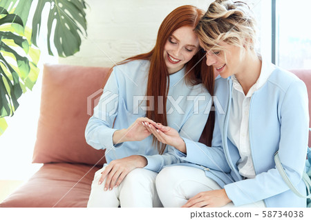 Two young caucasian women sit on sofa and show manicure Two young caucasian women sit on sofa and show manicure 58734038