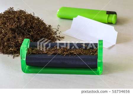 Green cigarette rolling machine, cigarette paper, green lighter and pile of tobacco on a white table Green cigarette rolling machine, cigarette paper, green lighter and pile of tobacco on a white table 58734656