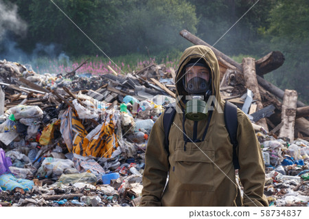 A working man in a gas mask against the backdrop of burning garbage. A lot of plastic bags thrown to the dump. From the plastic debris goes smoke. On the man, the khaki color encephalic suit 58734837