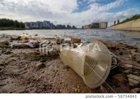 Disposable plastic cup on the city beach. The littered shore of the lake with plastic trash on the background of apartment buildings. Disposable plastic cup on the city beach. The littered shore of the lake with plastic trash on the background of apartment buildings. 58734843