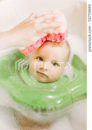 Baby swimming with green neck swim ring. Mom washing baby head with a red sponge Baby swimming with green neck swim ring. Mom washing baby head with a red sponge 58759668