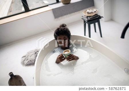 Curly dark-skinned woman drinking cappuccino while lying in bath Curly dark-skinned woman drinking cappuccino while lying in bath 58762871